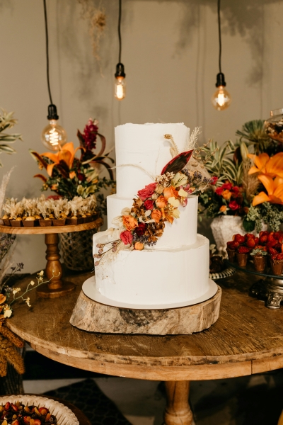 A three-tiered white wedding cake with a harvest colored floral design sits on a wooden table surrounded by other autumnal decor.