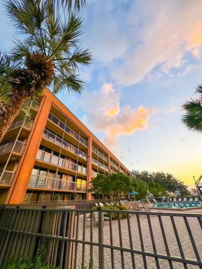 A rust-colored five-story building glows at sunset beside a pool and palm trees.  