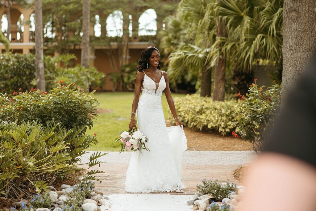 A beautiful bride appears at the end of an aisle outdoors, surrounded by gardens and palm trees. 