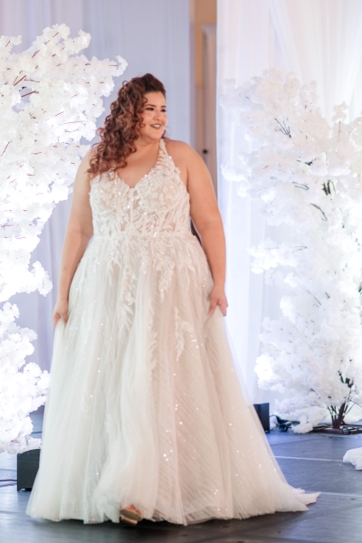 A beautiful bride wearing a halter A-line gown surrounded by tall white florals in a wedding expo fashion show.