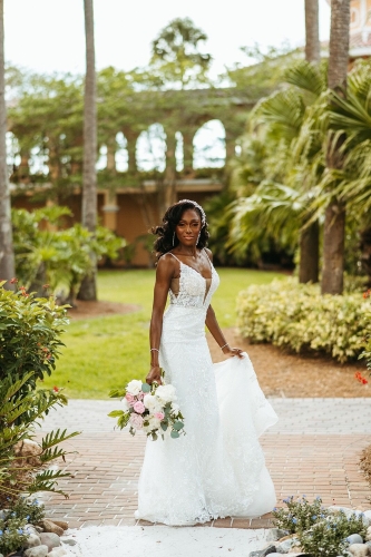 A woman in a lacy white gown holds a bouquet of pink and white roses while standing in a courtyard surrounded by palm trees. Your Orlando destination wedding could be full of lush palm trees and tropical foliage.