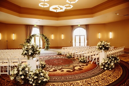 A warm ballroom space with a geometric chandelier set up with white chairs and white floral arrangements facing an altar decorated in white florals. 