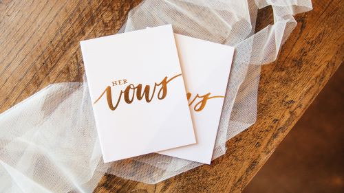 Two white cards with gold ink, one reading "Her Vows" and the other "His Vows" in calligraphy, resting on a piece of white tulle fabric against a wooden surface. 