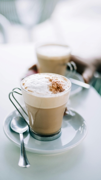 A close-up of a cappuccino on a white saucer sitting on a white table. A hand holds another coffee sits behind, out of focus. 