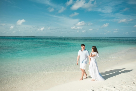 A couple dressed in white walk along a beach with clear blue water. A wedding registry can also include experiences and a honeymoon fund.