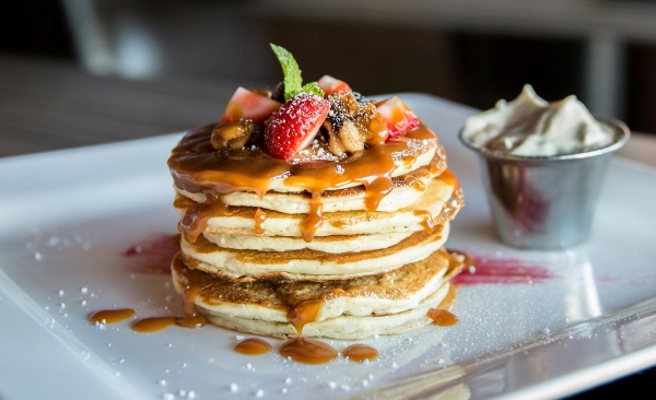 A stack of pancakes on a plate beside a small cup of whipped cream, topped with caramel, fruit, and a sprig of mint. 