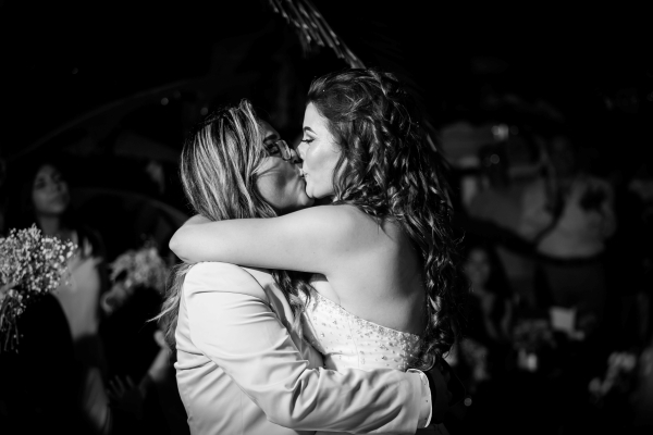 A black and white image of two women dressed in white kissing and embracing on the dance floor. 