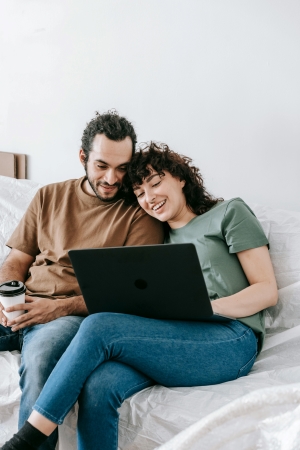 A couple sits on a white couch looking at a computer screen together. Making a wedding registry is a team effort, so be sure to work with your partner to create a registry you both love.