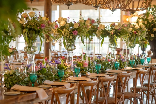 A long, wooden table with wooden chairs set with teal glasses and large floral centerpieces. 