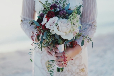 A woman in a long-sleeved white dress holds a bouquet of white roses with dark red flowers, red berries, and sprigs of eucalyptus. Dark maroons are a beautiful and classic color palette for a winter wedding.