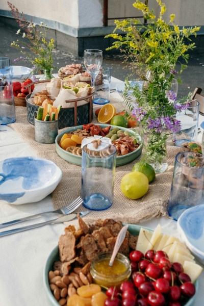 A tablescape with vases of yellow and purple wildflowers, a bowl with nuts, cherries, and cheese, a charcuterie plate, a burlap table runner, and blue and white ceramic dishes. 