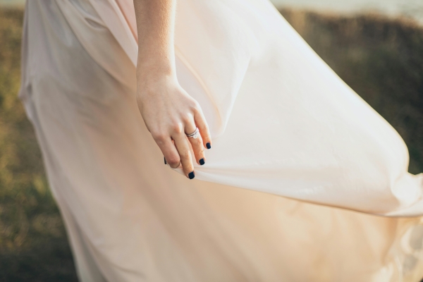 A close-up of a woman’s hand with black nail polish, an engagement ring, and a wedding band holding a flowing, lightweight white skirt. 