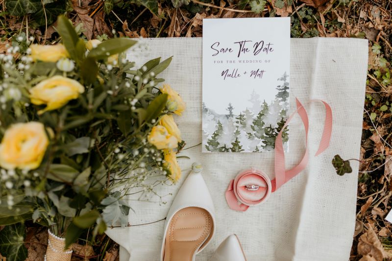 A beautiful flat lay featuring a bouquet of yellow flowers, white shoes, a wedding ring in a pink box, and a save-the-date postcard.  