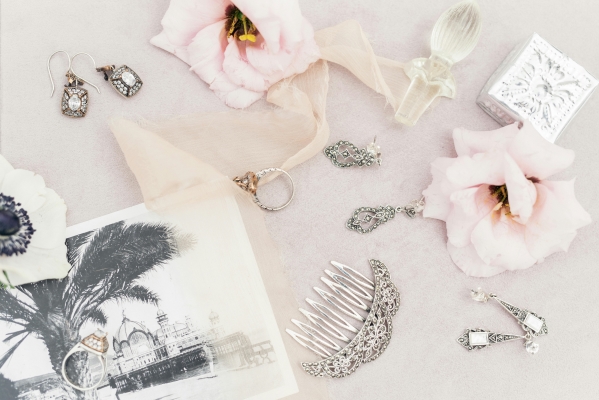 A flat lay photo featuring various pairs of antique earrings, a vintage comb, an old photograph, and several soft pink flowers. To honor lost loved ones at your wedding, wear a piece of their jewelry.