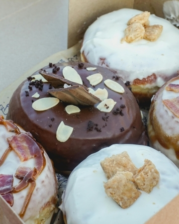 A close-up of donuts glazed in chocolate and white icing, topped with sliced almonds, cereal, and bacon. Donuts are a popular wedding dessert alternative.