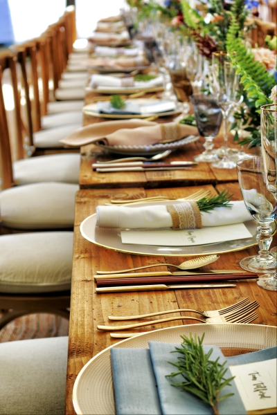 A close up of a wooden table and table settings with white linens, gold silverware, and sprigs of rosemary.