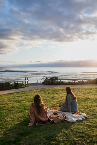 Two women on a picnic blanket sitting on a green lawn overlooking a body of water at sunset. Simply spending quality time together is a great option for a bridesmaid proposal. 