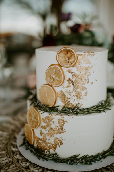 A two-tier wedding cake decorated with orange slices, gold leaf, and rosemary.