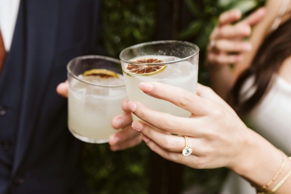 A man in a suit and a woman in a white dress wearing a diamond ring and wedding band toast cocktail glasses garnished with dried citrus slices.  