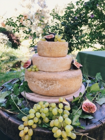 Three tiers of cheese wheels stacked like a cake, decorated with grapes and figs in an outdoor setting: a savory wedding dessert alternative. 