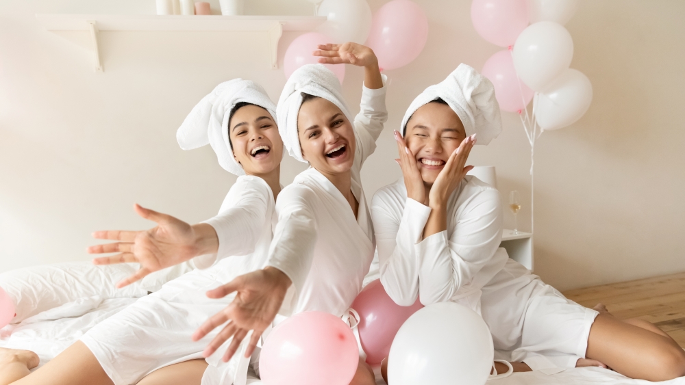 Three women in white robes and towel head wraps smile and pose surrounded by pink and white balloons. 
