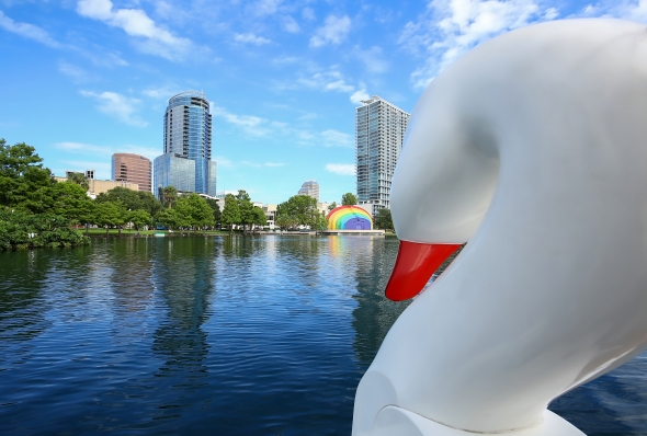 The back of a white Swan Boat looking out over Lake Eola with the city skyline and the rainbow-painted Walt Disney Amphitheater in the distance. 