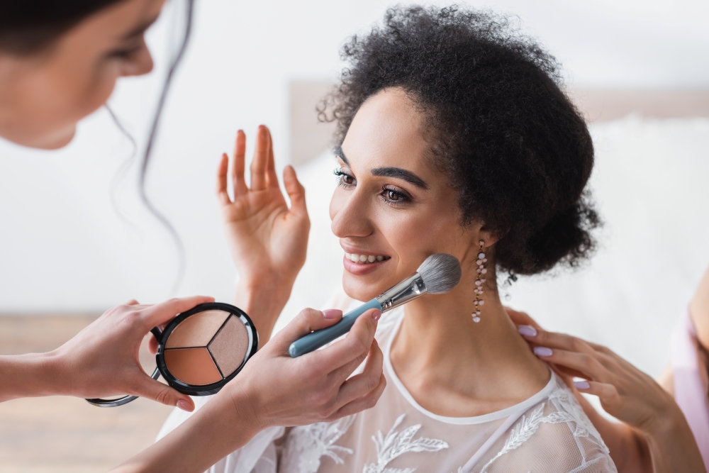 A bride in a white dress smiles while having makeup applied. 