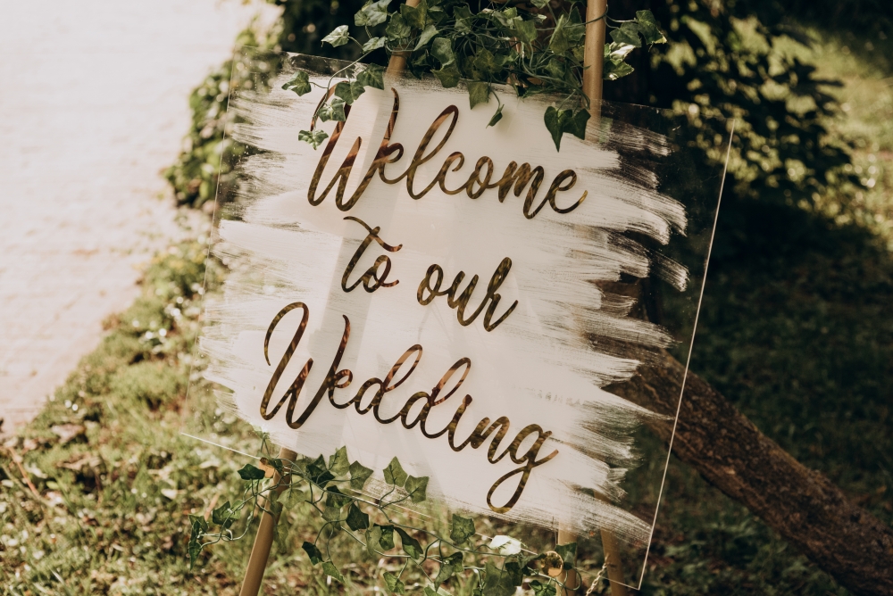 A clear acrylic sign with white paint and gold lettering that reads Welcome to Our Wedding standing on an outdoor easel. 