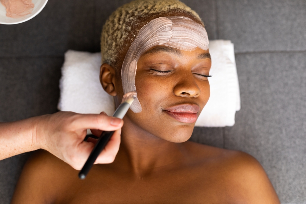 A woman smiles, relaxed, while lying on white towels as someone gives her a facial. 