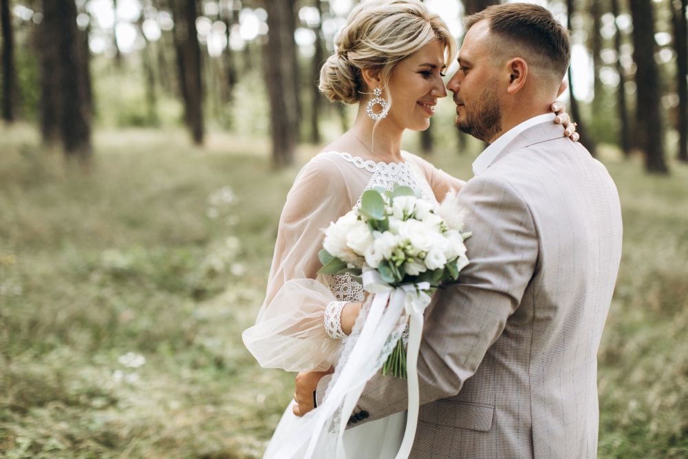 A bride in a white gown embraces a man in a light gray suit, carrying a small white bouquet and surrounded by trees.