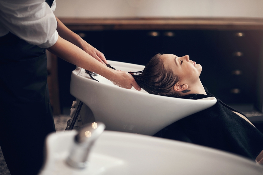 A woman lies back with her hair in a washbowl as a stylist washes her hair.