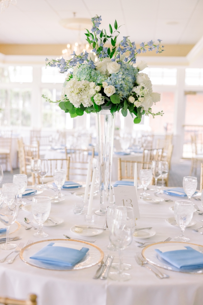 A tall floral arrangement on a table in a bright room, made of hydrangeas, greenery, and other blue florals.