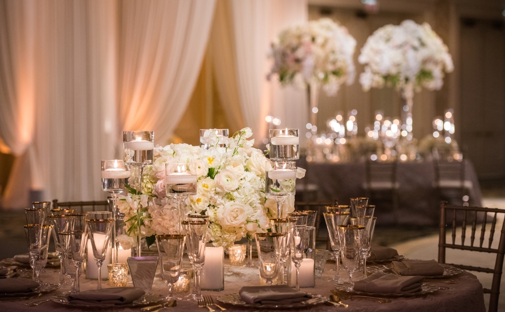 A ballroom wedding with tables set with candles, glassware, and beautiful white centerpieces, some high and some low.