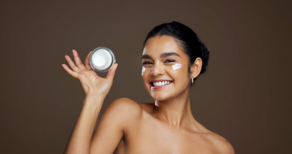 A bare-shouldered woman smiles at the camera holding a small container of skincare cream.