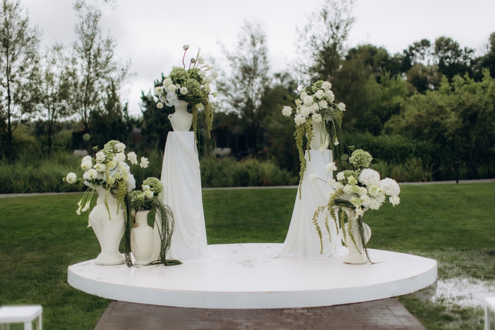 A round altar surrounded by grass and decorated with white vases on columns decorated in white and green florals.