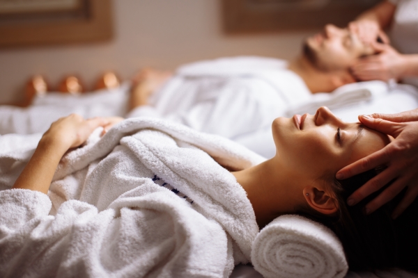 A man and a woman in white robes lie on their backs while hands massage their heads. 