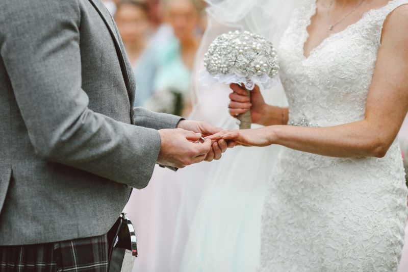 A close-up image of a bride and groom where the groom is putting the ring on the bride. The bride is wearing a formfitting white dress holding a bouquet of tulle and pearls. The groom is wearing a grey blazer and appears to be wearing a kilt. 