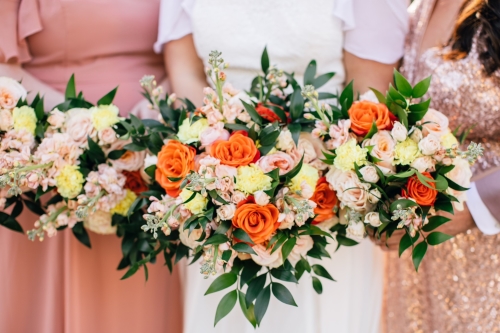 A closeup of bouquet with orange, pink, and yellow flowers held by two bridesmaids in blush pink dresses and a bride. Ask your besties to stand by your side with a unique bridesmaid proposal.