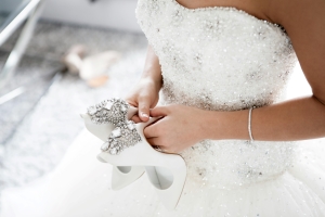 Bride wearing a strapless wedding dress decorated in beads while holding a pair of white high heels.